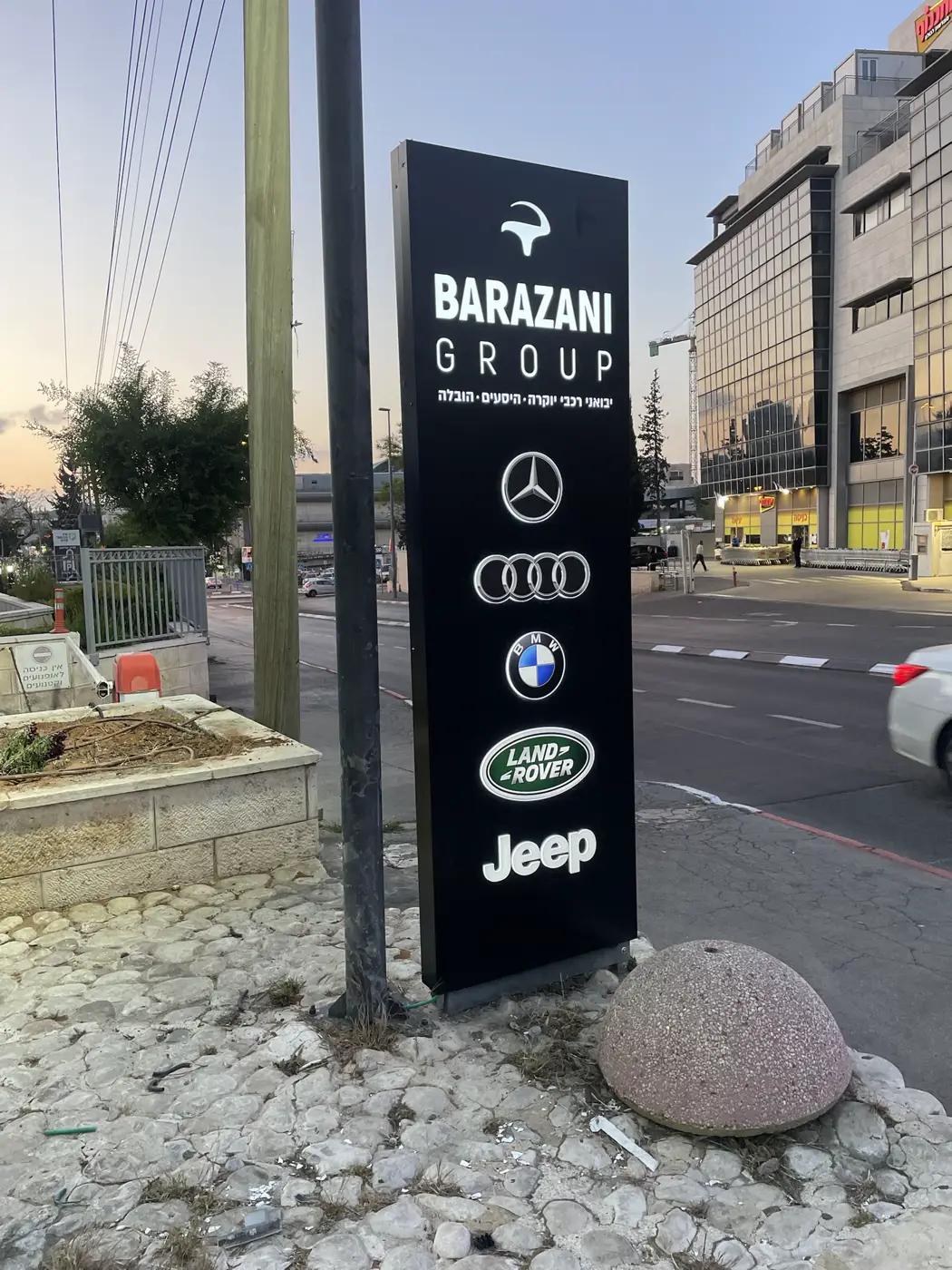 טוטם עומד שחור (קאט אאוט) ליד כביש עם לוגו החברה ולוגואים של מותגי רכב מוארים, בשעת ערב