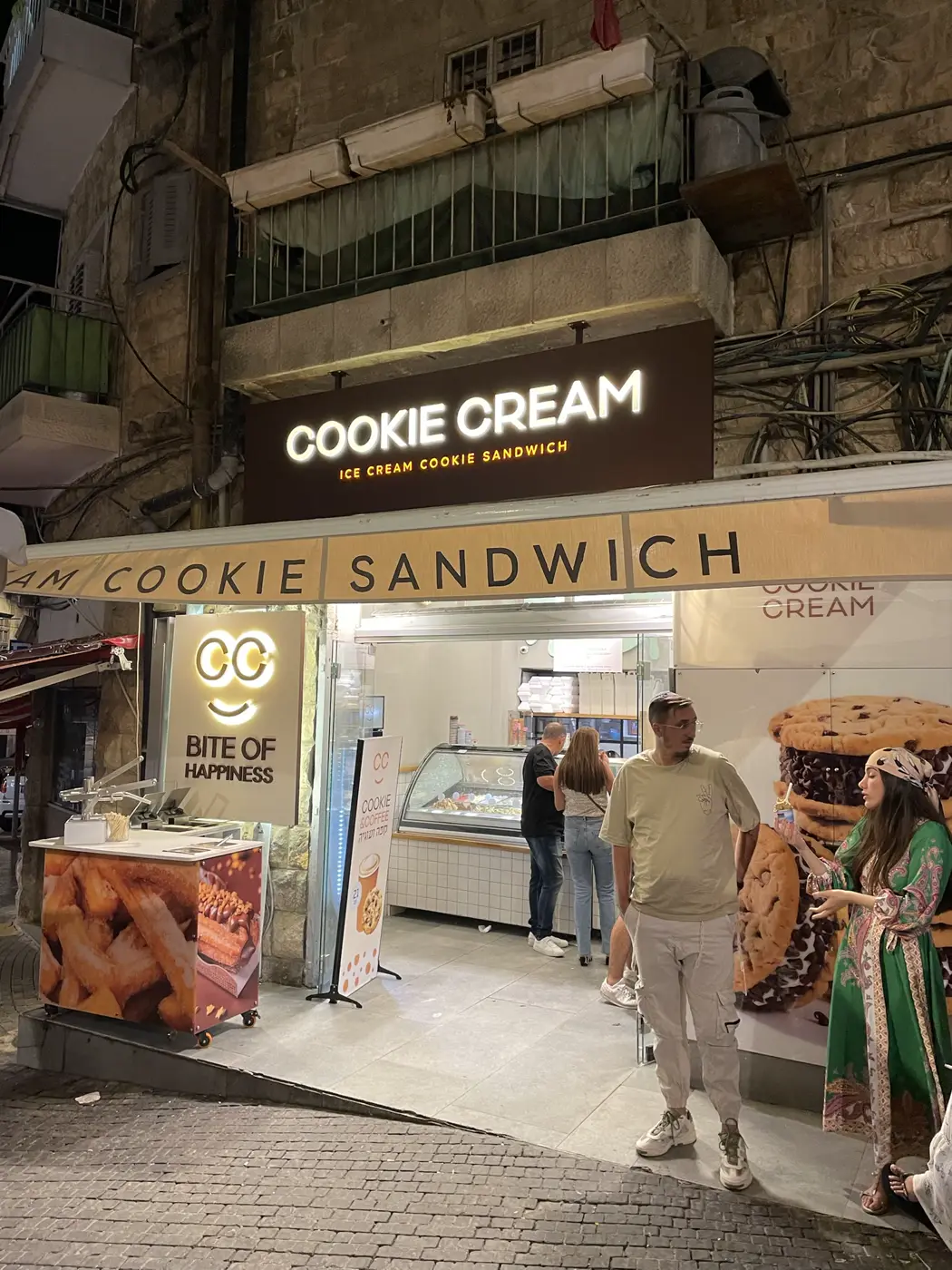 ארגז קאט אאוט חום תלוי על בניין אבן מעל גלידריה, אותיות לבנות ושורה כתומה מוארות בלילה, ושלט לוגו נוסף בצד