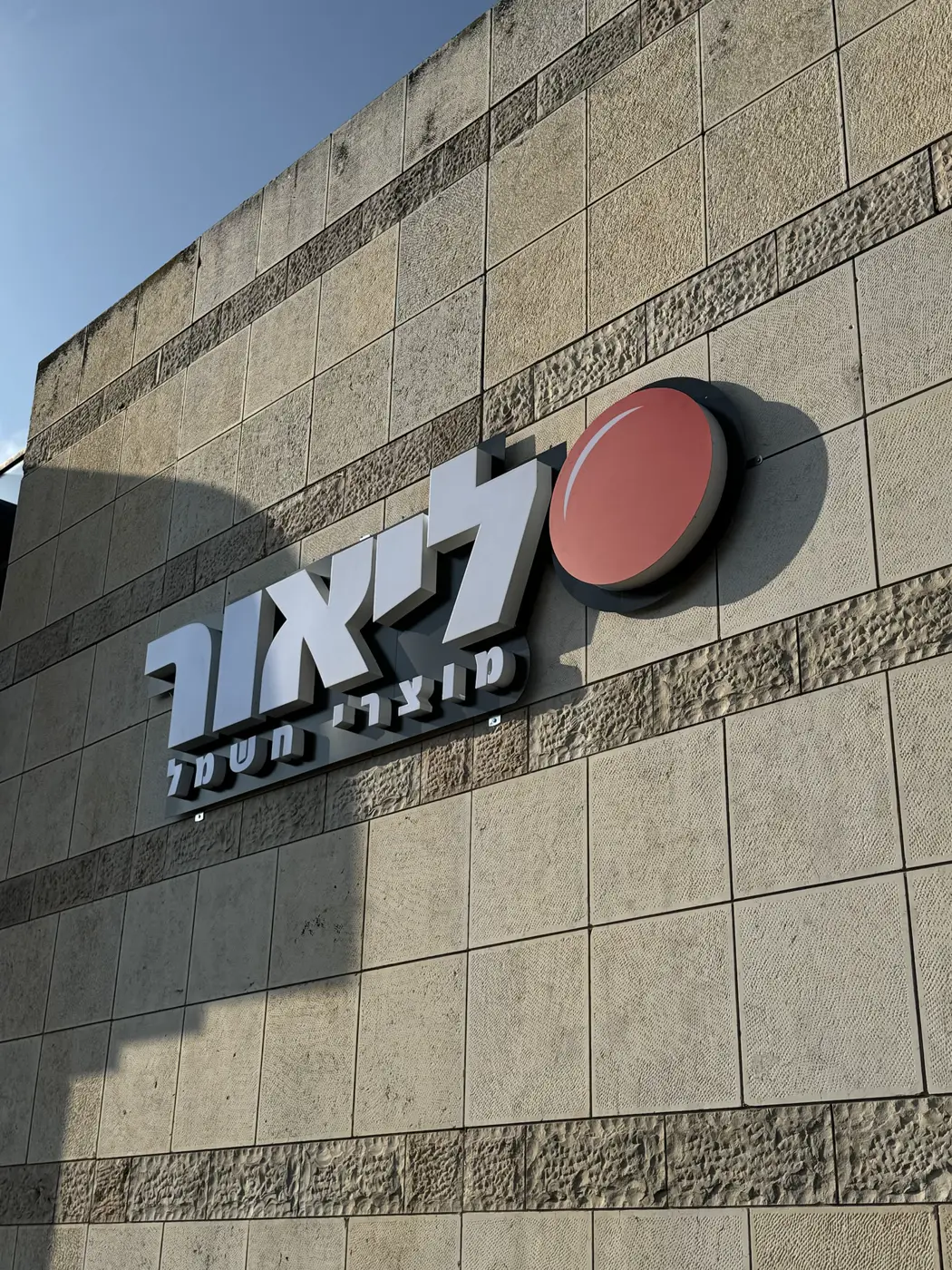 אותיות עבריות לבנות בנויות על פאנל אפור ולוגו עגול אדום בולט, על קיר אבן של חנות