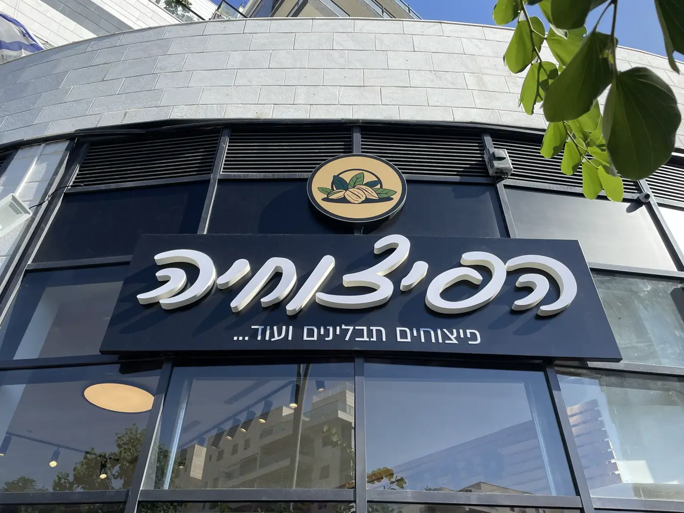 אותיות עבריות בנויות לבנות בכתב יד על פאנל שחור, ומעליו לוגו עגול מואר, בחזית חנות פיצוחים