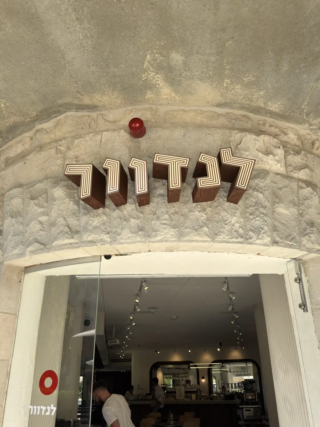 אותיות עבריות בנויות עמוקות עם חזית מוארת בדוגמת פסים ודופן בגימור חלודה, מעל קשת אבן בכניסה לבית קפה