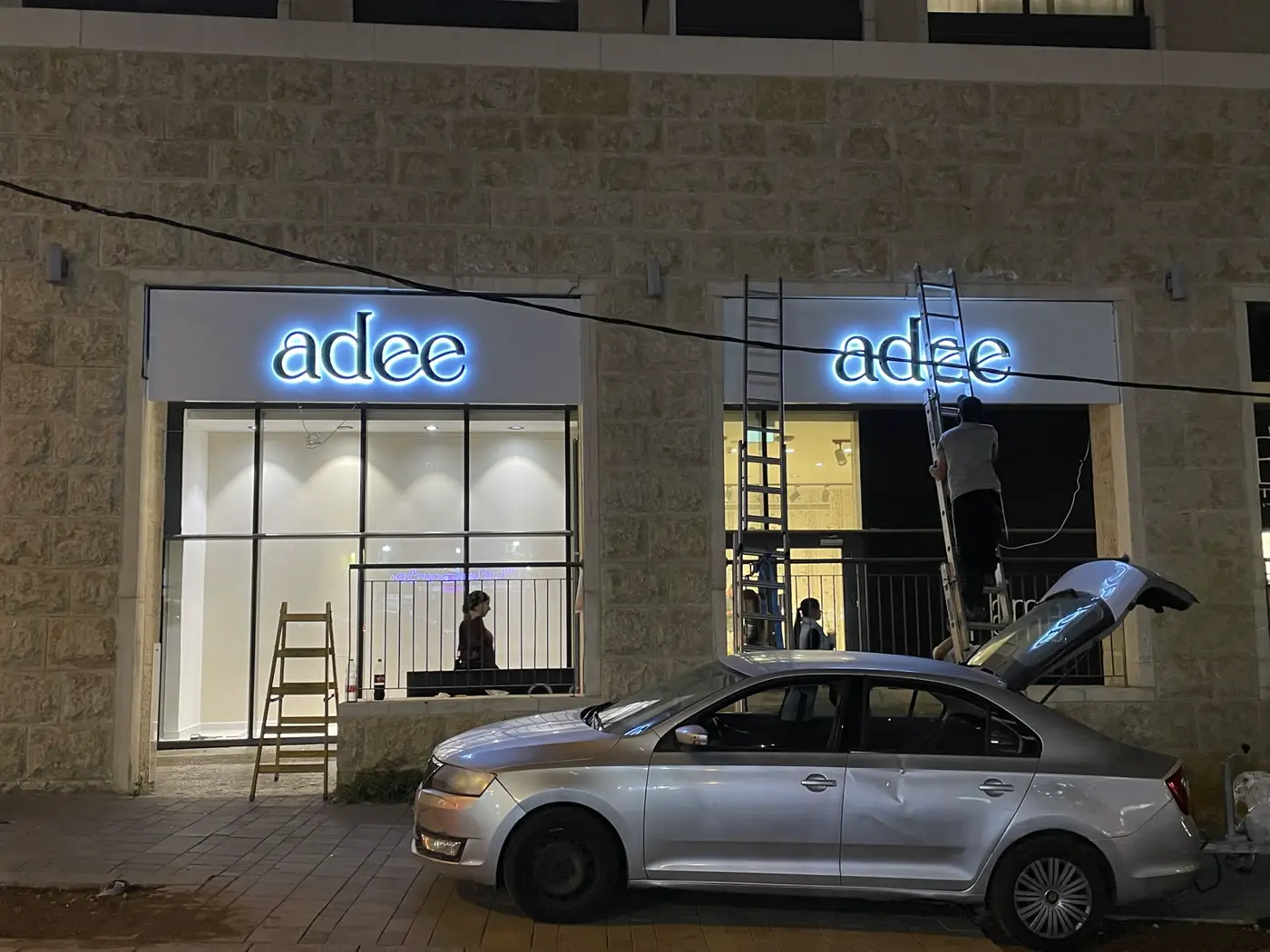 שתי חזיתות של חנות: אותיות שחורות עם הילה לבנה-כחלחלה על פאנל לבן מעל ויטרינות, בזמן התקנה בלילה