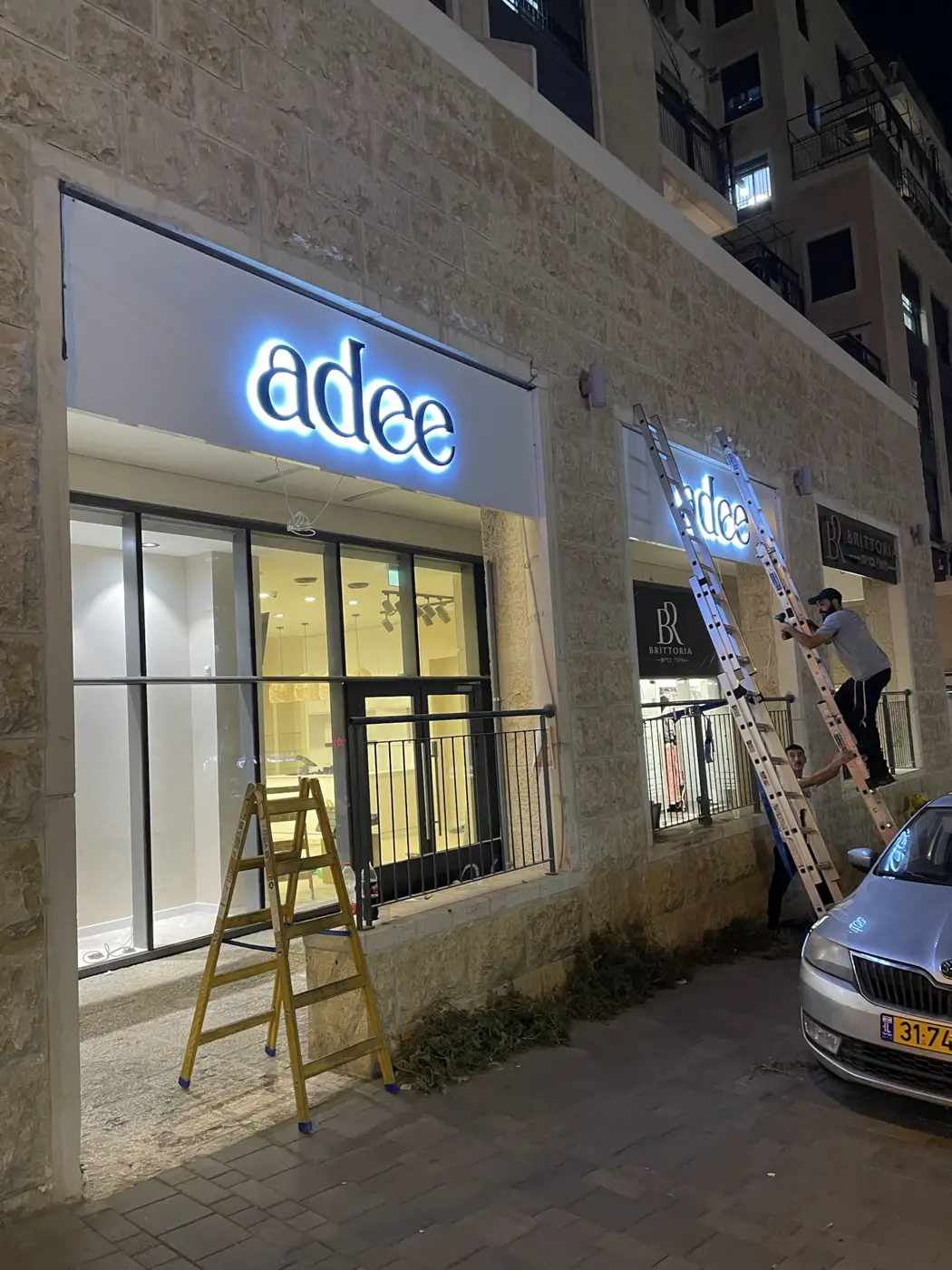 התקנה של אותיות עם הילה לבנה על פאנל לבן מעל ויטרינה בחזית אבן, צוות על סולם