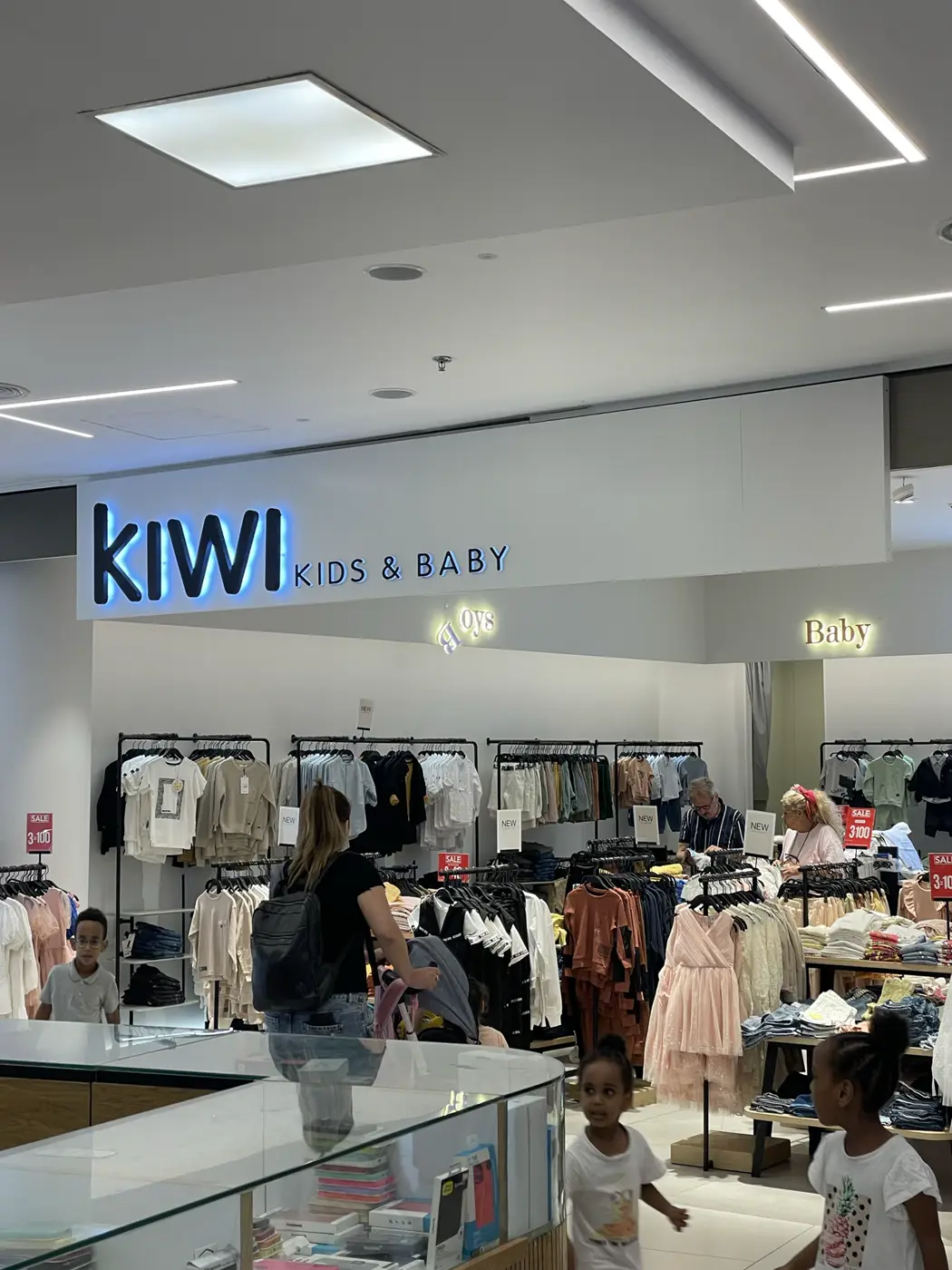 אותיות כהות עם הילה כחולה מעל כניסה של חנות בגדי ילדים בקניון, ושלטי מחלקות קטנים מוארים בפנים