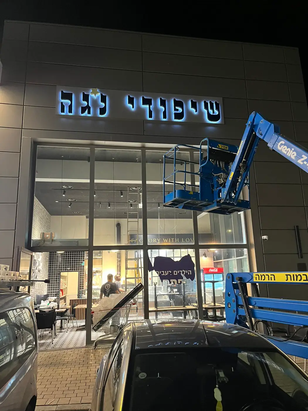 אותיות עבריות גדולות שחורות עם הילה כחולה על חזית מסעדה בחיפוי אלומיניום, בזמן התקנה עם במת הרמה