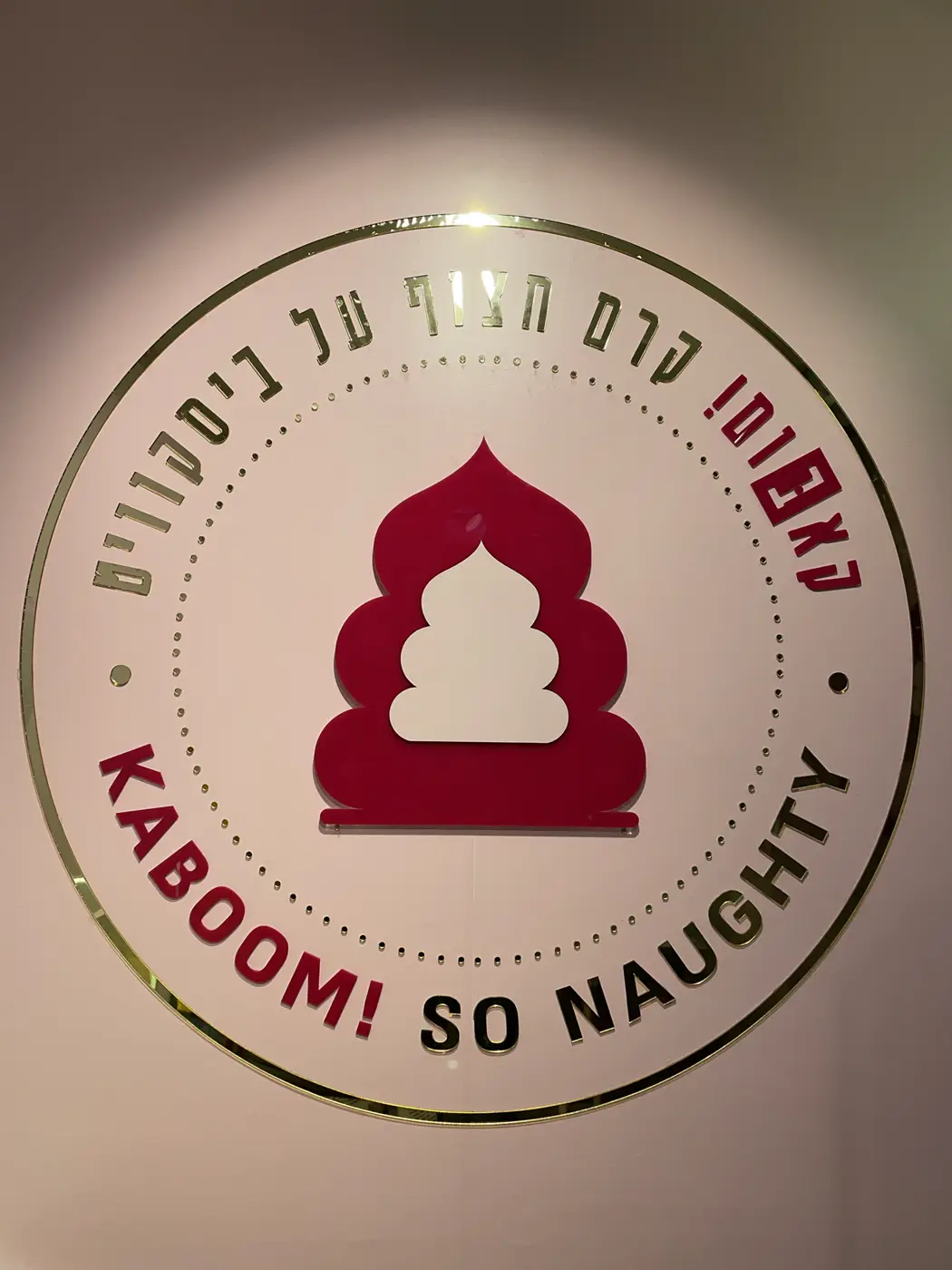 לוגו עגול גדול על קיר ורוד: טבעת ונקודות מראה זהב, אייקון בורדו-לבן ואותיות בעברית ובאנגלית