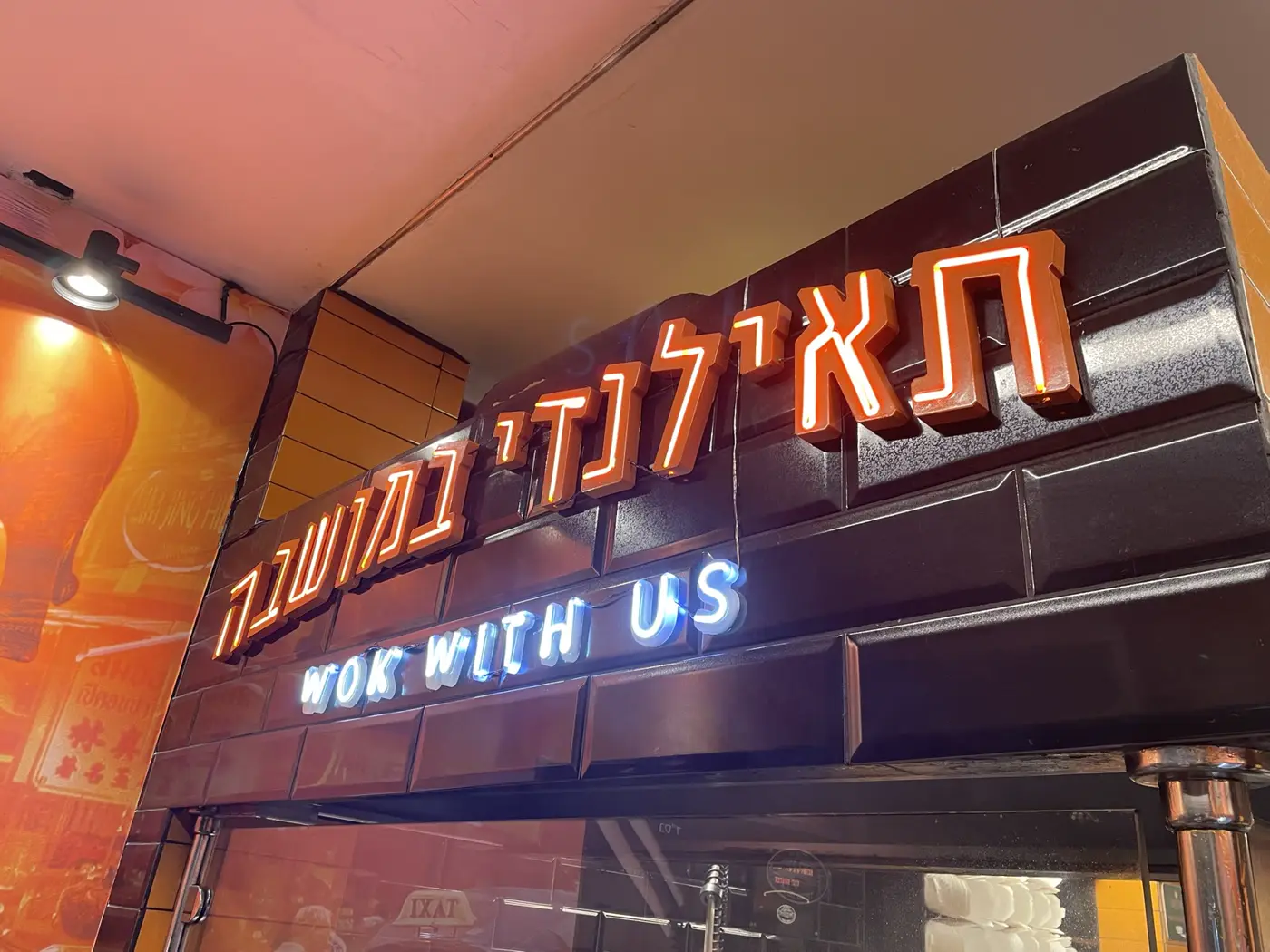 אותיות עבריות בנויות כתומות עם ניאון בתוכן וטקסט באנגלית בניאון לבן, מעל כניסה למסעדה תאילנדית